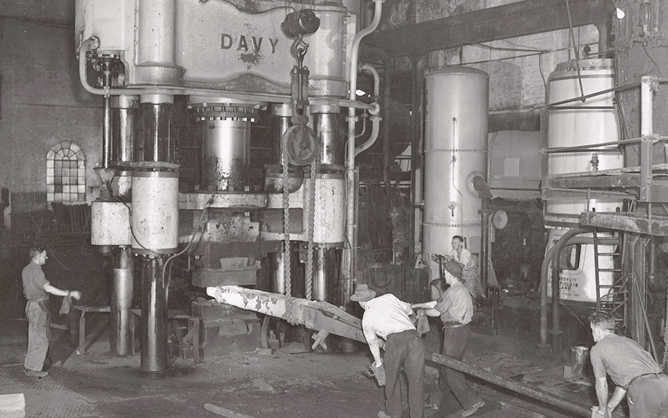 Davy Press in operation. Workers using billet holder to move heated billet under press, crane with pulley and chains attached to hold billet above, operator on left, hydraulic reservoir and steam intensifier visible in right of image.